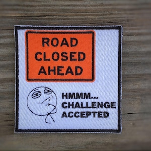 以下が含まれることがあります： 黒い文字と漫画の顔が付いた白いパッチ。テキストは「Road Closed Ahead」と「Hmmm... Challenge Accepted」です。