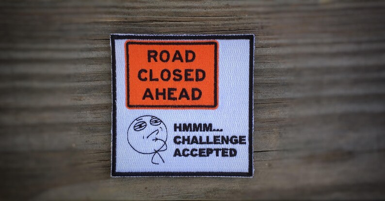 Op de afbeelding: Een witte patch met zwarte tekst en een cartoon gezicht. De tekst luidt "Road Closed Ahead" en "Hmmm... Challenge Accepted".