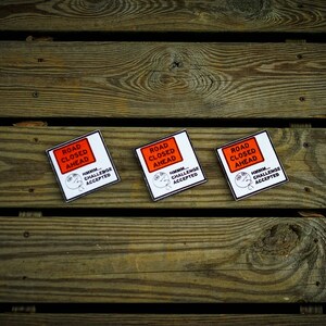 Op de afbeelding: Drie witte vierkante stickers met zwarte tekst en een rood rechthoek met de tekst "ROAD CLOSED AHEAD". De tekst onder de rechthoek zegt "HUMAN CHALLENGE ACCEPTED".