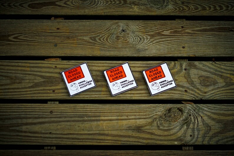 Op de afbeelding: Drie witte vierkante stickers met zwarte tekst en een rood rechthoek met de tekst "ROAD CLOSED AHEAD". De tekst onder de rechthoek zegt "HUMAN CHALLENGE ACCEPTED".