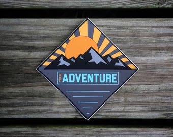 May include: A diamond-shaped sticker with a mountain range and a sun rising behind it. The sticker has a blue and gray background with the text "Your Adventure" in a light blue font.