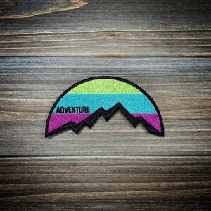 May include: A colorful embroidered patch with a mountain range design and the word "ADVENTURE" in black text. The patch is a half-circle shape with a green, blue, and purple color scheme.