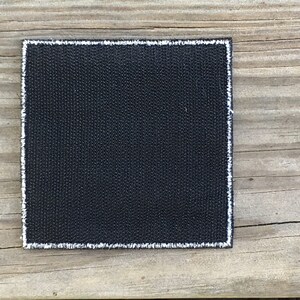 Op de afbeelding: Een zwarte vierkante patch met witte stiksels rond de randen. De patch is gemaakt van een geweven stof.