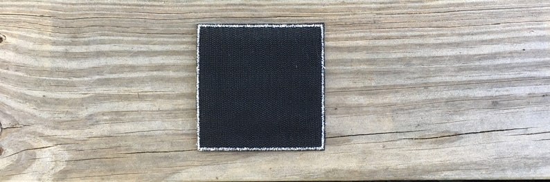 Op de afbeelding: Een zwarte vierkante patch met witte stiksels rond de randen. De patch is gemaakt van een geweven stof.