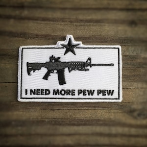 Puede incluir: Parche blanco con una silueta negra de un rifle y el texto "I NEED MORE PEW PEW" en negro. Una pequeña estrella está en la esquina superior izquierda.