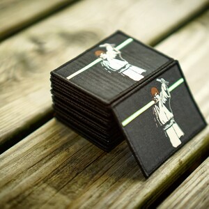 May include: A stack of black fabric patches with a white graphic of a person in a white robe holding a lightsaber. The patches are on a wooden surface.