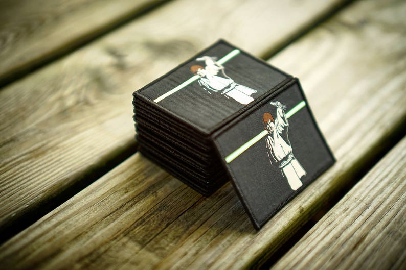 May include: A stack of black fabric patches with a white graphic of a person in a white robe holding a lightsaber. The patches are on a wooden surface.