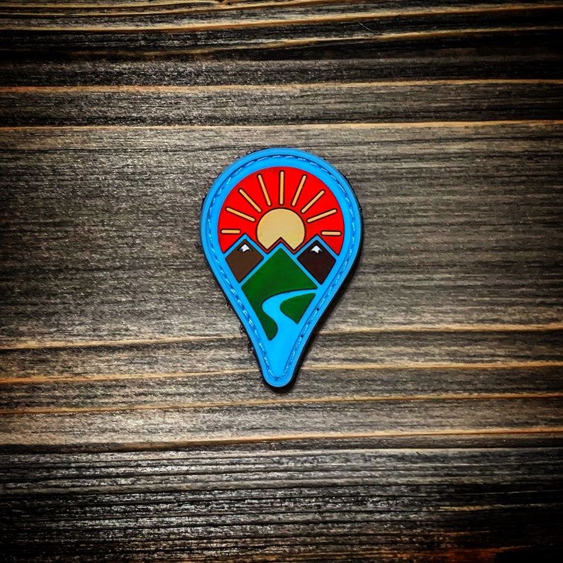 May include: A blue and red embroidered patch shaped like a map pin with a sun, mountains, and a river inside.