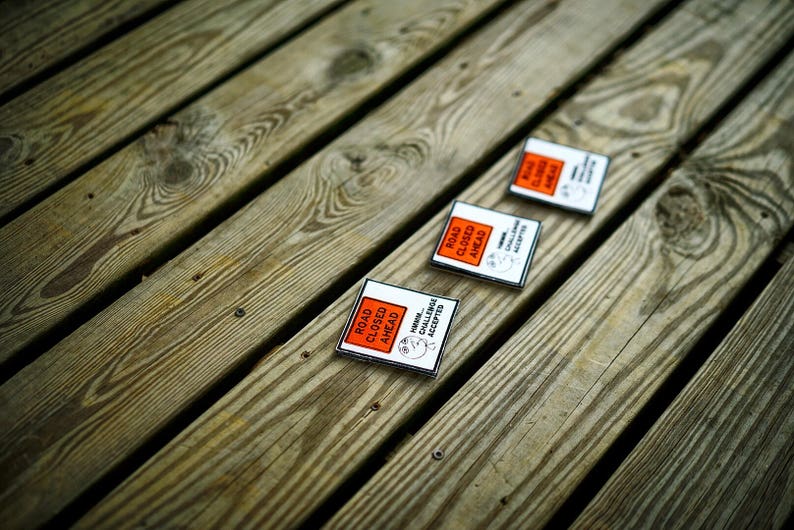 Op de afbeelding: Drie oranje en zwarte borden met de tekst 'Weg afgesloten' met een cartoonfiguur dat de uitdaging aanneemt. De borden liggen op een houten oppervlak.