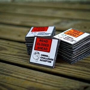 Op de afbeelding: Een stapel van vier witte en oranje borden met de tekst "Weg afgesloten" met een zwart-wit meme-beeld van een persoon die "Hmmm... Uitdaging geaccepteerd" zegt.