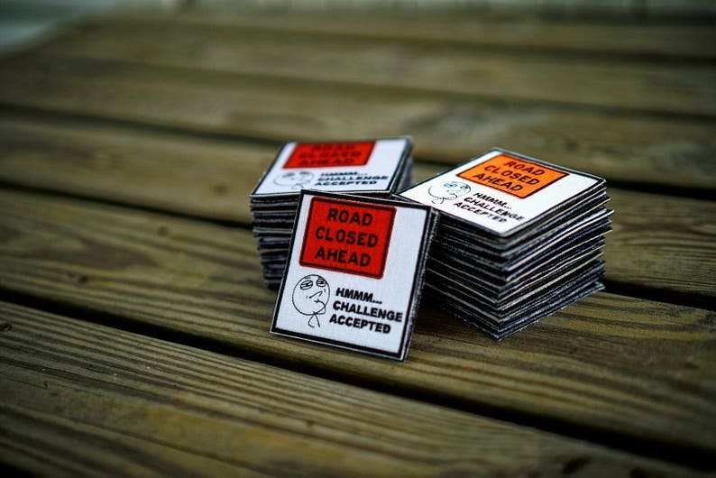 Op de afbeelding: Een stapel van vier witte en oranje borden met de tekst "Weg afgesloten" met een zwart-wit meme-beeld van een persoon die "Hmmm... Uitdaging geaccepteerd" zegt.