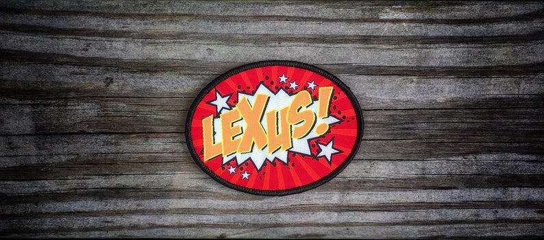 May include: A round, red and black patch with a white and yellow comic book style explosion graphic. The text "LEXUS!" is written in yellow letters.