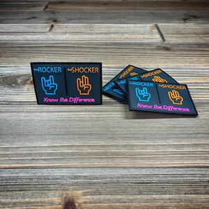 K&ouml;nnte beinhalten: F&uuml;nf schwarze Patches mit dem Text "the ROCKER" und "the SHOCKER" in Orange und Blau. Jeder Patch hat eine andere Handgeste mit dem Text "Know the Difference" in Pink.