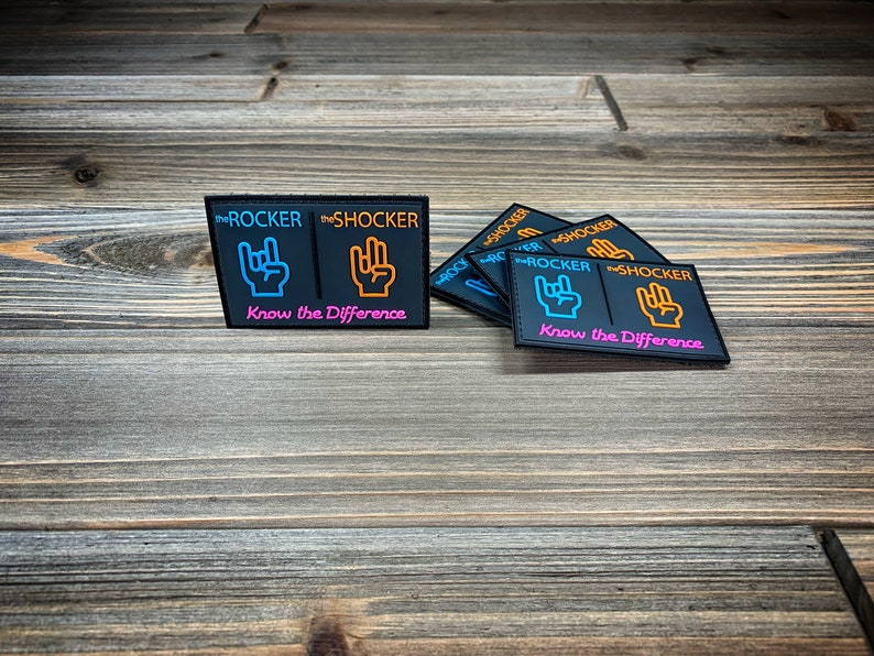 K&ouml;nnte beinhalten: F&uuml;nf schwarze Patches mit dem Text "the ROCKER" und "the SHOCKER" in Orange und Blau. Jeder Patch hat eine andere Handgeste mit dem Text "Know the Difference" in Pink.