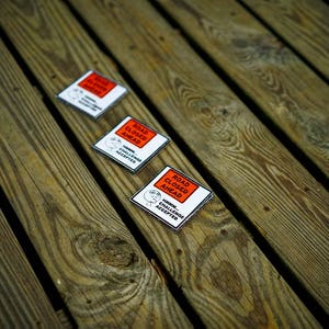 Op de afbeelding: Drie witte en rode stickers met de tekst "ROAD CLOSED AHEAD" en "KILLER CHALLENGE ACCEPTED" op een houten oppervlak.