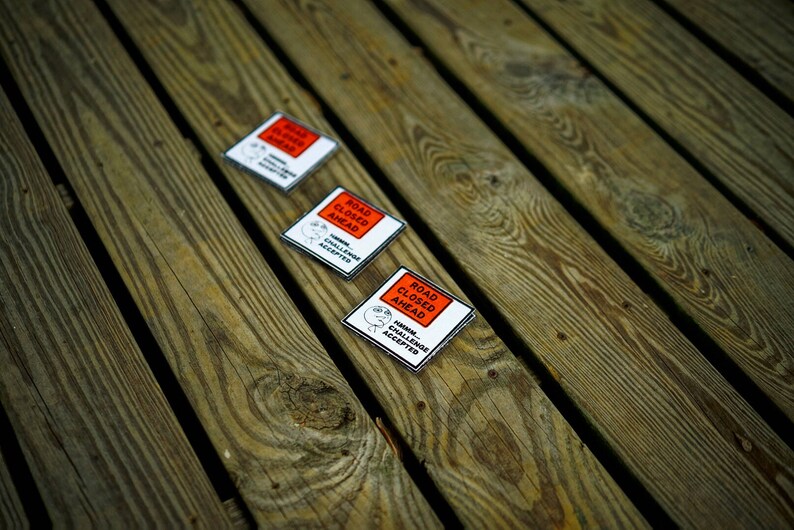 Op de afbeelding: Drie witte en rode stickers met de tekst "ROAD CLOSED AHEAD" en "KILLER CHALLENGE ACCEPTED" op een houten oppervlak.