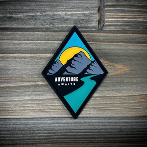 May include: A black diamond-shaped patch with a blue sky, yellow sun, grey mountains, and a turquoise river. The text "Adventure Awaits" is printed in white.