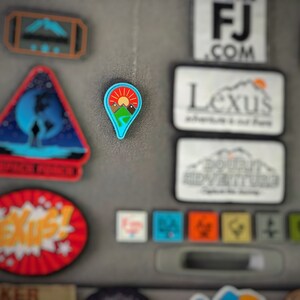 May include: A blue and green pin with a mountain range and a red sun. The pin is shaped like a location marker.