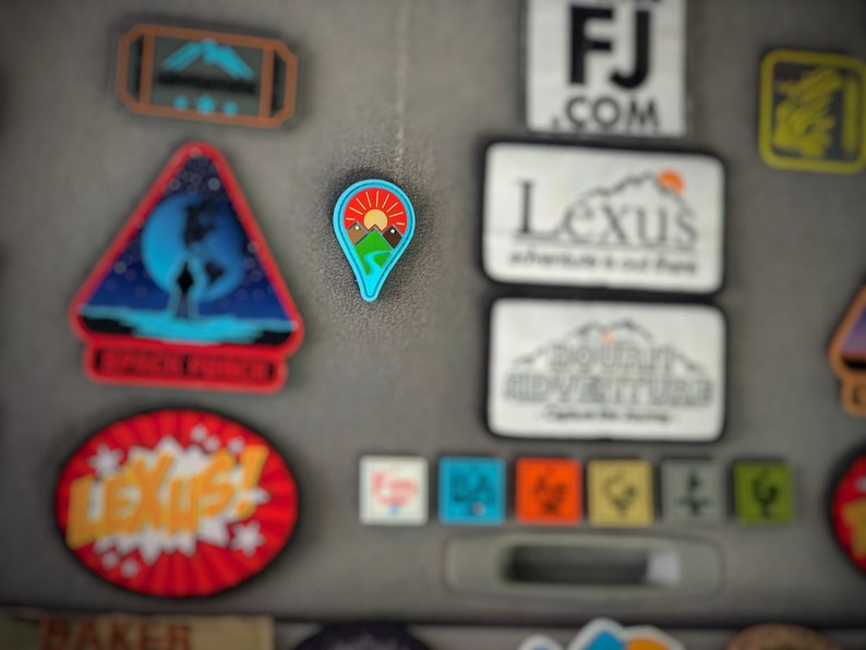 May include: A blue and green pin with a mountain range and a red sun. The pin is shaped like a location marker.