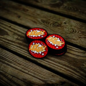 May include: Three round, red and yellow patches with the word "Lexus!" in a comic book style font. The patches are on a wooden surface.