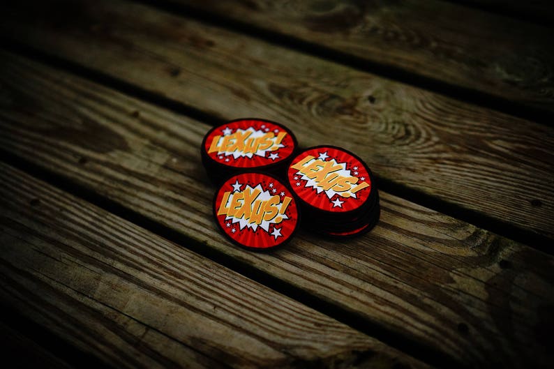 May include: Three round, red and yellow patches with the word "Lexus!" in a comic book style font. The patches are on a wooden surface.