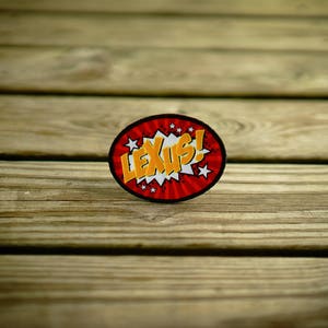 May include: A red and white circle sticker with the word "LEXUS!" in yellow letters and white stars around the edge. The sticker is on a wooden surface.
