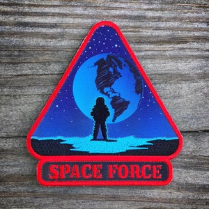 May include: A blue and black triangular patch with a red border. The patch features a silhouette of an astronaut standing in front of a blue and white Earth. The text "SPACE FORCE" is printed in white on a black banner at the bottom of the patch.