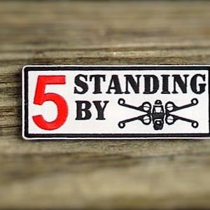 May include: A white rectangular patch with black text that reads "5 Standing By" and a black and white graphic of a star wars X-wing fighter.