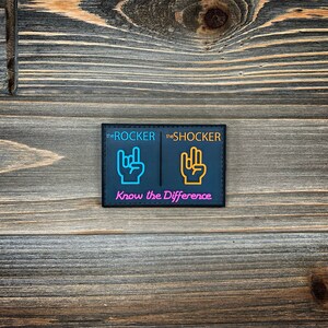 K&ouml;nnte beinhalten: Schwarzer Patch mit dem Text "the ROCKER" und "the SHOCKER" in Wei&szlig;. Jede Seite zeigt eine Handgeste in einer anderen Farbe. Die linke Seite hat eine blaue Hand mit ausgestrecktem Zeige- und kleinem Finger. Die rechte Seite hat eine gelbe Hand mit ausgestrecktem Zeige-, Mittel- und kleinem Finger. Der Text "Know the Difference" ist unten in Pink gedruckt.