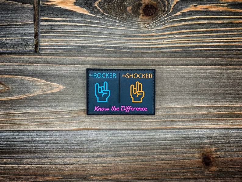 K&ouml;nnte beinhalten: Schwarzer Patch mit dem Text "the ROCKER" und "the SHOCKER" in Wei&szlig;. Jede Seite zeigt eine Handgeste in einer anderen Farbe. Die linke Seite hat eine blaue Hand mit ausgestrecktem Zeige- und kleinem Finger. Die rechte Seite hat eine gelbe Hand mit ausgestrecktem Zeige-, Mittel- und kleinem Finger. Der Text "Know the Difference" ist unten in Pink gedruckt.