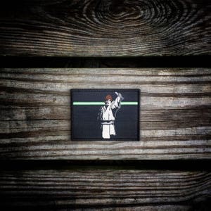 May include: Black and green embroidered patch featuring a white silhouette of a person holding a lightsaber. The patch has a green line across the top.