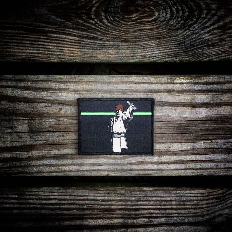 May include: Black and green embroidered patch featuring a white silhouette of a person holding a lightsaber. The patch has a green line across the top.