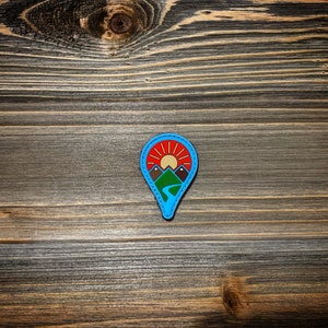 May include: A blue and red pin with a mountain range and a sun design. The pin is on a wooden surface.