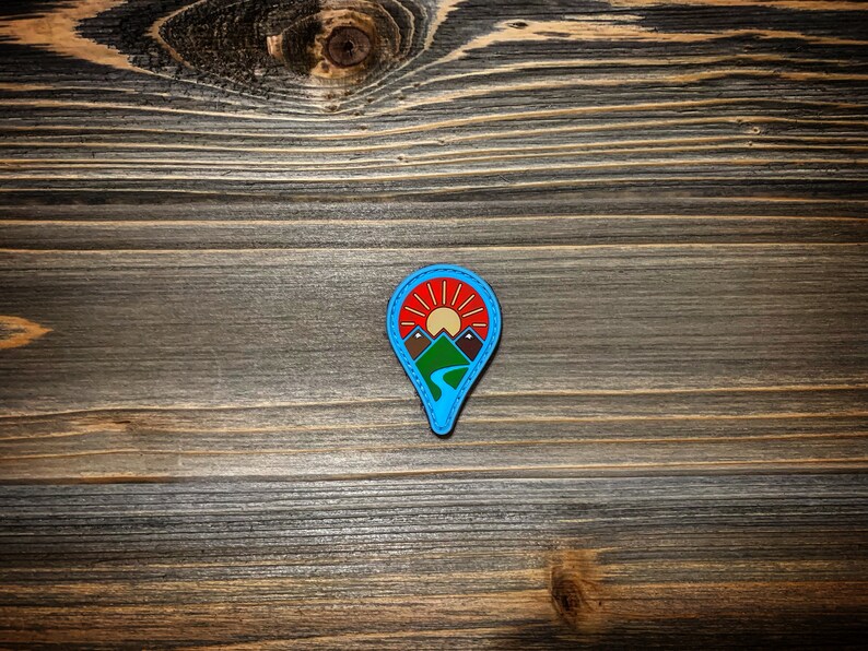 May include: A blue and red pin with a mountain range and a sun design. The pin is on a wooden surface.