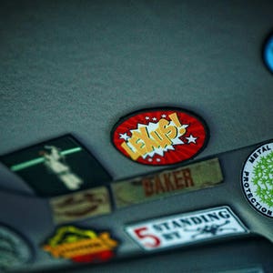 May include: A close-up of a car's ceiling with various colorful patches and stickers. The stickers feature different designs, including a red and yellow sticker with the word "LEXUS!" in a comic book style font, a green and black sticker with the text "METAL TECH PROTECTION EVOLVED" and a white sticker with the text "STANDING".