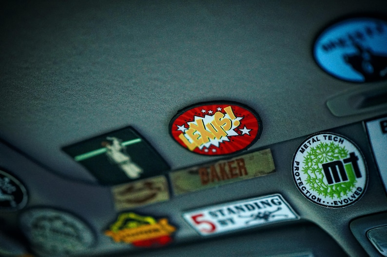 May include: A close-up of a car's ceiling with various colorful patches and stickers. The stickers feature different designs, including a red and yellow sticker with the word "LEXUS!" in a comic book style font, a green and black sticker with the text "METAL TECH PROTECTION EVOLVED" and a white sticker with the text "STANDING".