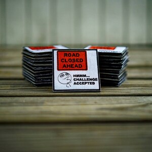 Op de afbeelding: Een witte vierkante patch met zwarte tekst die "Road Closed Ahead" luidt en een zwart-witte illustratie van een persoon met de tekst "Hmmm... Challenge Accepted". De patch ligt bovenop een stapel vergelijkbare patches.