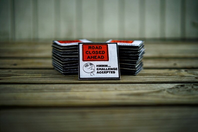 Op de afbeelding: Een witte vierkante patch met zwarte tekst die "Road Closed Ahead" luidt en een zwart-witte illustratie van een persoon met de tekst "Hmmm... Challenge Accepted". De patch ligt bovenop een stapel vergelijkbare patches.
