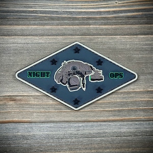 May include: A dark blue diamond-shaped patch with white stitching. The patch features a black and white illustration of a person wearing a night vision helmet and a green inscription that reads "NIGHT OPS".