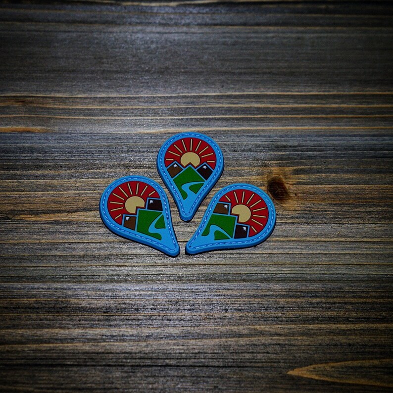 May include: Three blue, red, and brown felt patches shaped like teardrops with a mountain scene and a sun inside each patch.