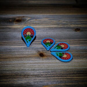 May include: Four blue and red pinback buttons with a mountain and sun design. The buttons are arranged in a circle on a wooden surface.