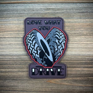 Op de afbeelding: Een grijze en zwarte patch met een hartvormig ontwerp gemaakt van twee bandenprofielen. De tekst "LOVE WHAT YOU DRIVE" is op de patch gedrukt.