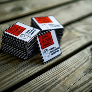 Op de afbeelding: Een stapel vierkante stickers in rood en wit met de tekst "Road Closed Ahead" en een cartoon gezicht met de tekst "Hmm... Challenge Accepted".