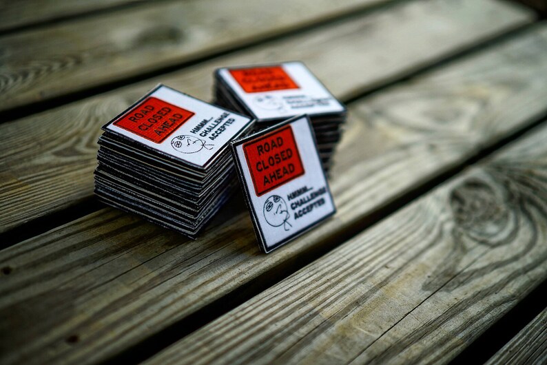 Op de afbeelding: Een stapel vierkante stickers in rood en wit met de tekst "Road Closed Ahead" en een cartoon gezicht met de tekst "Hmm... Challenge Accepted".