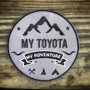 May include: A gray embroidered patch with a mountain range design, the text "MY TOYOTA MY ADVENTURE", and icons of an axe, a campfire, and a tent.
