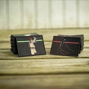 May include: Two black fabric patches with embroidered images of Star Wars characters. The left patch shows Luke Skywalker holding a lightsaber with a green blade. The right patch shows Darth Vader holding a lightsaber with a red blade.