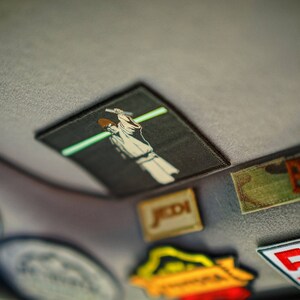 May include: A collection of colourful patches, including a Star Wars themed patch with a character wielding a lightsaber, a patch with the word "BAKER" in white letters on a green background, and a patch with the text "STAND BY" and the number "5" in white on a red background.