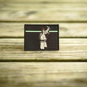 May include: A brown fabric patch with a green line across the top. The patch features a black and white illustration of a person holding a lightsaber.