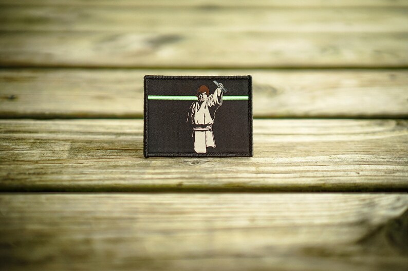 May include: A brown fabric patch with a green line across the top. The patch features a black and white illustration of a person holding a lightsaber.