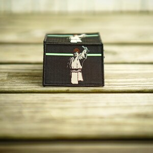 May include: A black and green embroidered patch featuring a character holding a lightsaber. The patch has a green line across the top and a white background.
